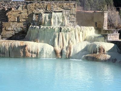 A tiered, man-made travertine waterfall structure with mineral deposits cascading into a turquoise pool, surrounded by stone retaining walls.