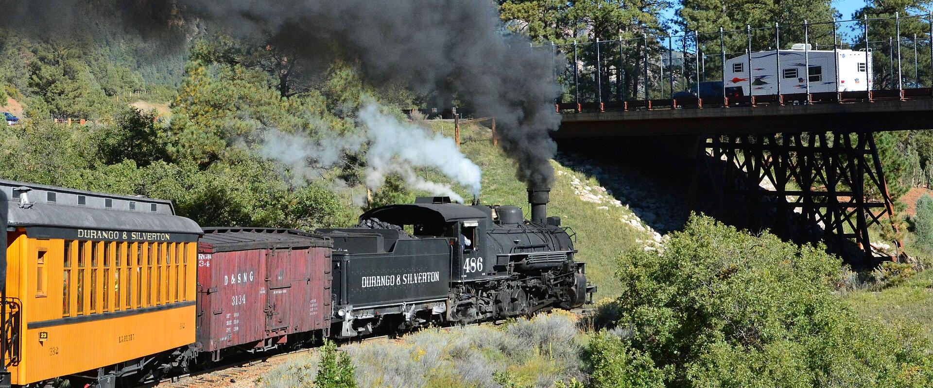 A black steam locomotive numbered 486 pulling a yellow passenger car and a red freight car along a track through a forested mountain landscape, with a trestle bridge visible in the background.