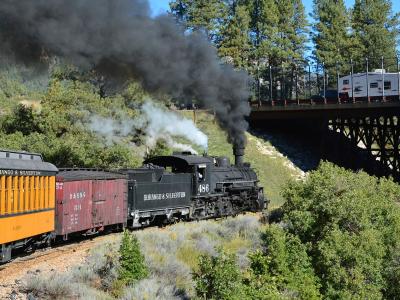A black steam locomotive numbered 486 pulling a yellow passenger car and a red freight car along a track through a forested mountain landscape, with a trestle bridge visible in the background.