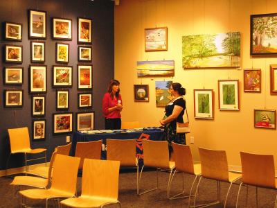 An indoor art gallery space with framed paintings on the walls, two women standing near a table, and rows of empty chairs facing the display area.