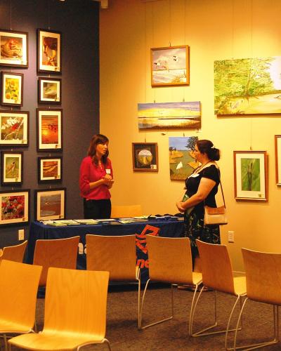 An indoor art gallery space with framed paintings on the walls, two women standing near a table, and rows of empty chairs facing the display area.