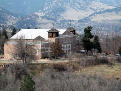 A large, distinctive building with a white roof and brownish walls, featuring multiple towers and arched windows, situated on a hillside with mountains in the background.
