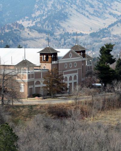 A large, distinctive building with a white roof and brownish walls, featuring multiple towers and arched windows, situated on a hillside with mountains in the background.