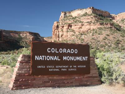 A large brown wooden sign reading "COLORADO NATIONAL MONUMENT" mounted on a brick base, with red rock cliffs and desert vegetation in the background.
