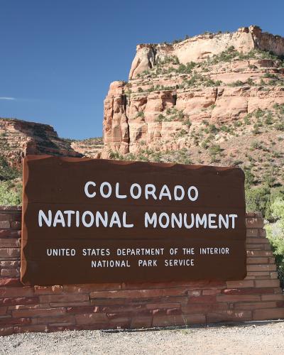 A large brown wooden sign reading "COLORADO NATIONAL MONUMENT" mounted on a brick base, with red rock cliffs and desert vegetation in the background.