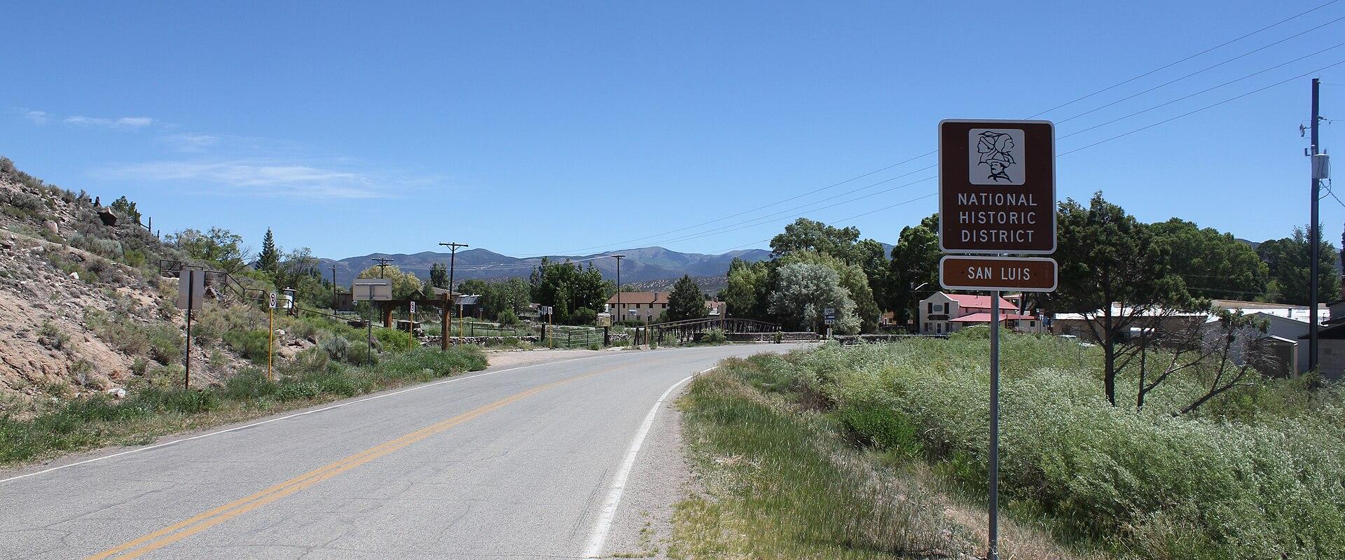 A roadside view of a paved road curving past a brown sign reading "NATIONAL HISTORIC DISTRICT" and "SAN LUIS", with mountains in the background and buildings visible in the distance.