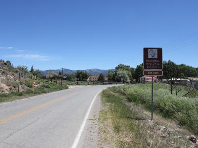 A roadside view of a paved road curving past a brown sign reading "NATIONAL HISTORIC DISTRICT" and "SAN LUIS", with mountains in the background and buildings visible in the distance.