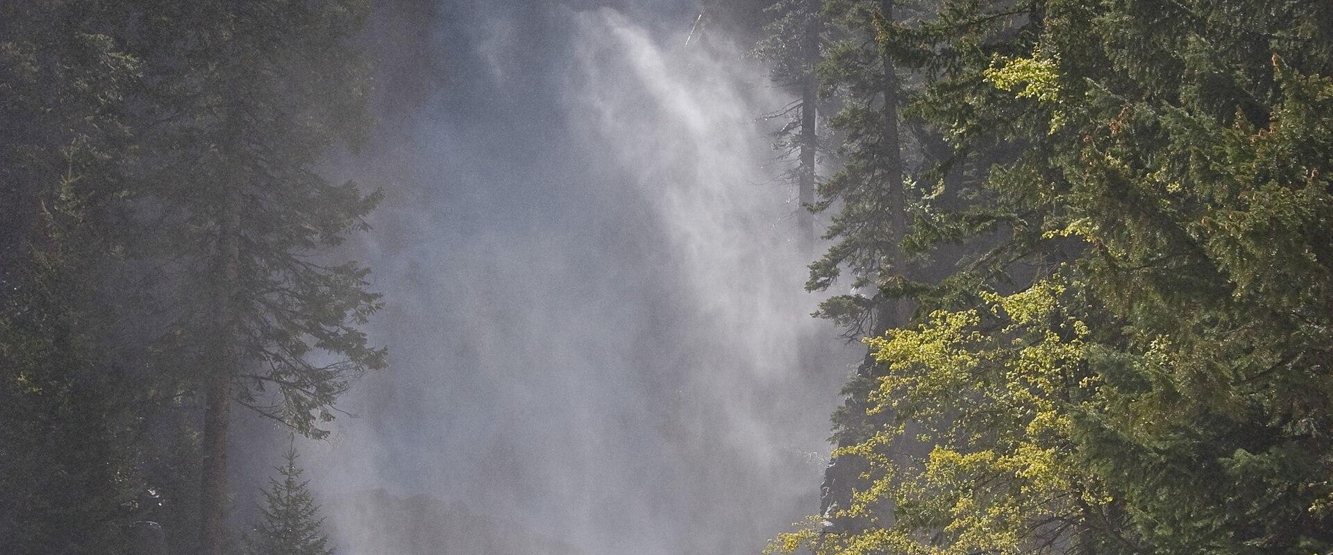A high-volume waterfall cascading down a rocky cliff face into a turbulent river below, surrounded by dense coniferous forest and featuring a small wooden footbridge crossing the rapids in the foreground.