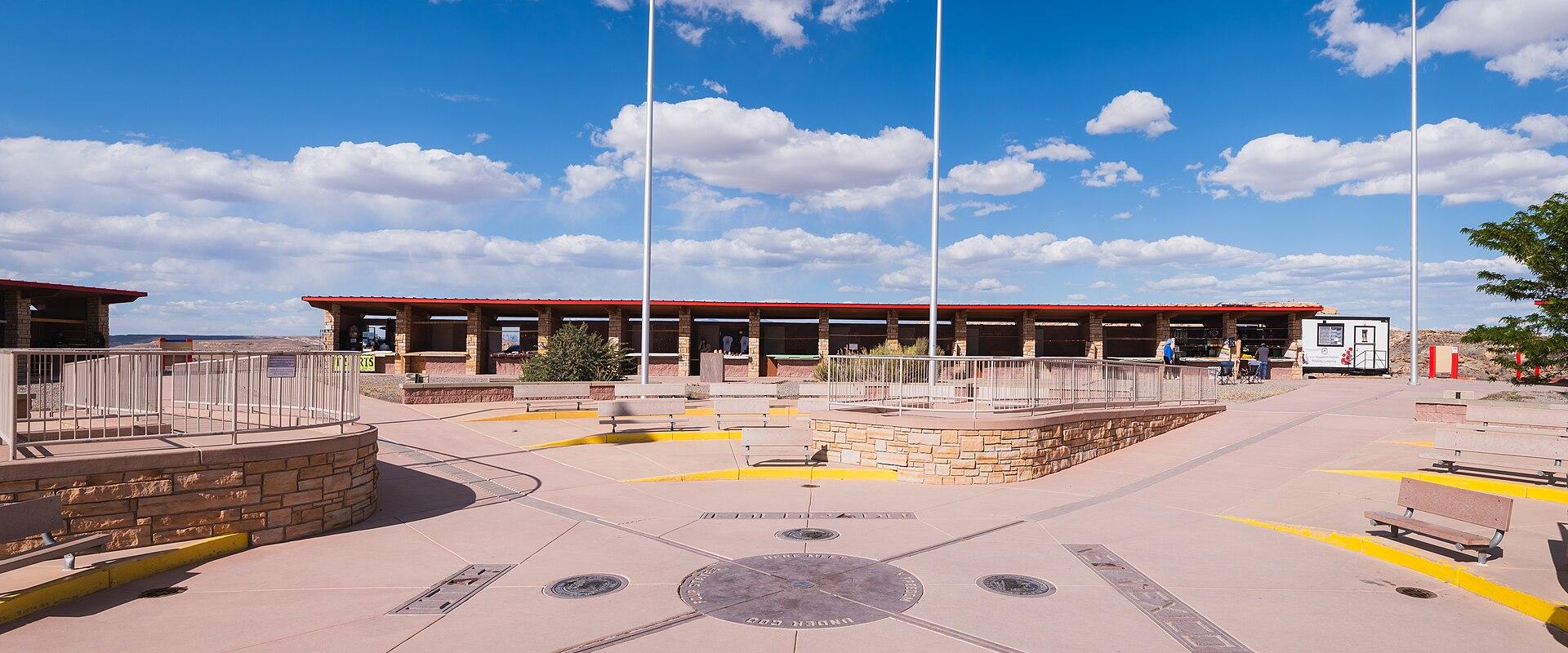A wide-angle view of the Four Corners Monument plaza showing the concrete ground with state names and seals, three flagpoles flying the US, Texas, and Navajo flags, and a visitor center building in the background.
