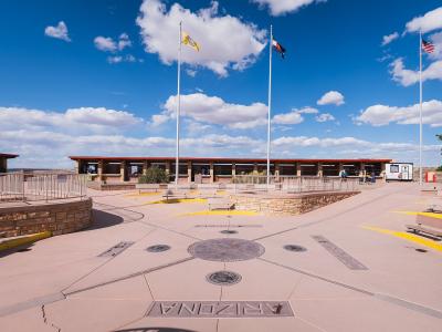 A wide-angle view of the Four Corners Monument plaza showing the concrete ground with state names and seals, three flagpoles flying the US, Texas, and Navajo flags, and a visitor center building in the background.