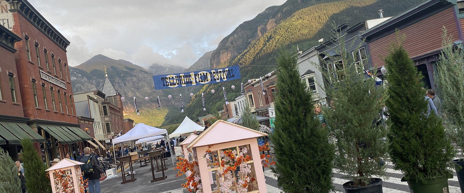 A street scene in a mountain town with a blue banner reading "TELLURIDE FILM FESTIVAL" strung across the road, flanked by historic buildings and mountains in the background.
