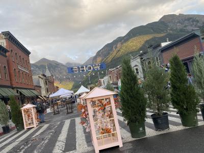 A street scene in a mountain town with a blue banner reading "TELLURIDE FILM FESTIVAL" strung across the road, flanked by historic buildings and mountains in the background.