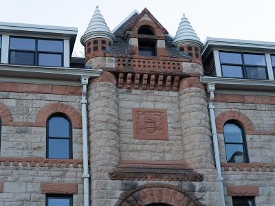 A close-up architectural view of the upper facade of a stone building featuring a central gable with two small conical turrets and a carved stone plaque displaying a crest and the date "1891".