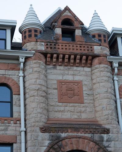 A close-up architectural view of the upper facade of a stone building featuring a central gable with two small conical turrets and a carved stone plaque displaying a crest and the date "1891".