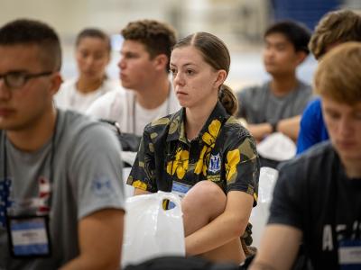 A group of young people, likely high school students, are seated in rows indoors; the central figure is a female cadet in a dark blue uniform with yellow floral patterns and a name tag, while others around her wear casual t-shirts.