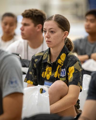 A group of young people, likely high school students, are seated in rows indoors; the central figure is a female cadet in a dark blue uniform with yellow floral patterns and a name tag, while others around her wear casual t-shirts.