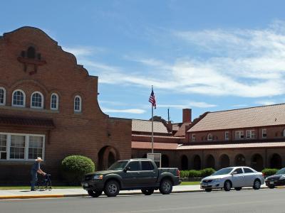 A wide-angle street view of a large, red-brick institutional building featuring distinctive Spanish Colonial Revival architecture, including a stepped parapet, arched windows, and a tiled roof, with an American flag flying in front and cars parked al
