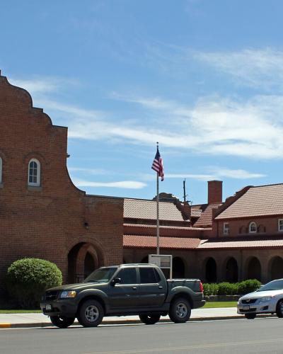 A wide-angle street view of a large, red-brick institutional building featuring distinctive Spanish Colonial Revival architecture, including a stepped parapet, arched windows, and a tiled roof, with an American flag flying in front and cars parked al