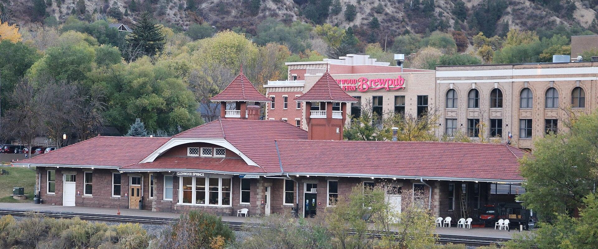 A brick train station building with a red tile roof and two small towers, situated next to train tracks with a hillside in the background.