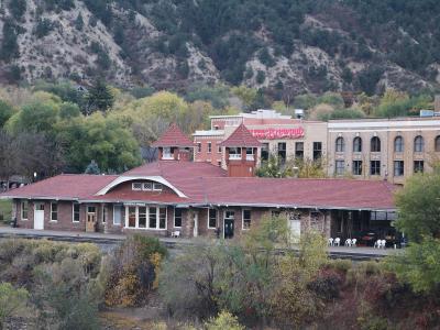 A brick train station building with a red tile roof and two small towers, situated next to train tracks with a hillside in the background.