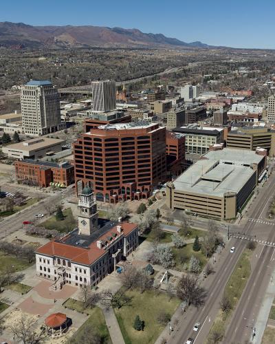 An aerial view of a downtown city center featuring a prominent historic building with a clock tower (El Paso County Courthouse) in the foreground, surrounded by mid-rise office buildings and a wide boulevard, with a backdrop of foothills and mountain