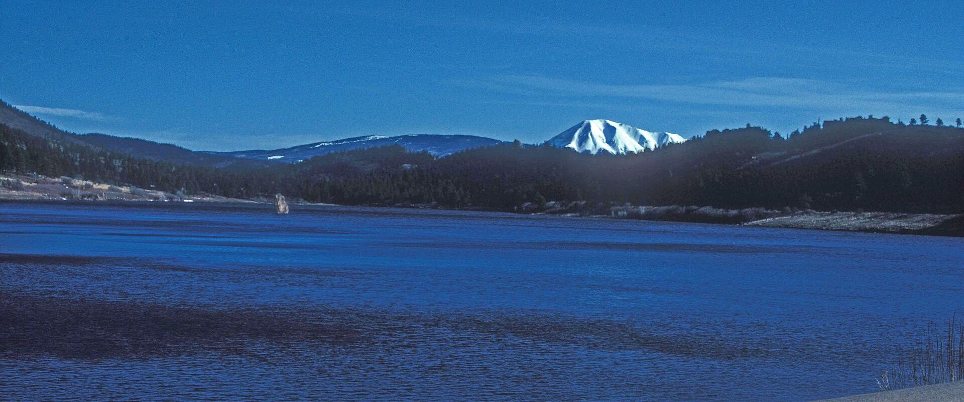 A wide blue lake with a stone retaining wall in the foreground and a snow-capped mountain peak visible in the distance under a clear blue sky.