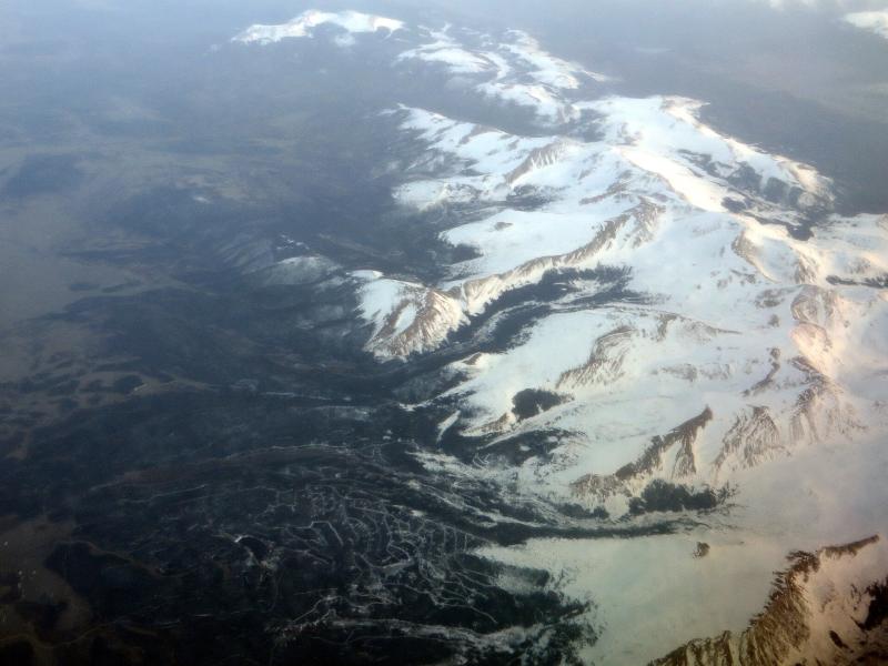 An aerial photograph of a snow-covered mountain range with a large ski resort visible in the foreground, featuring numerous groomed runs and lift infrastructure.