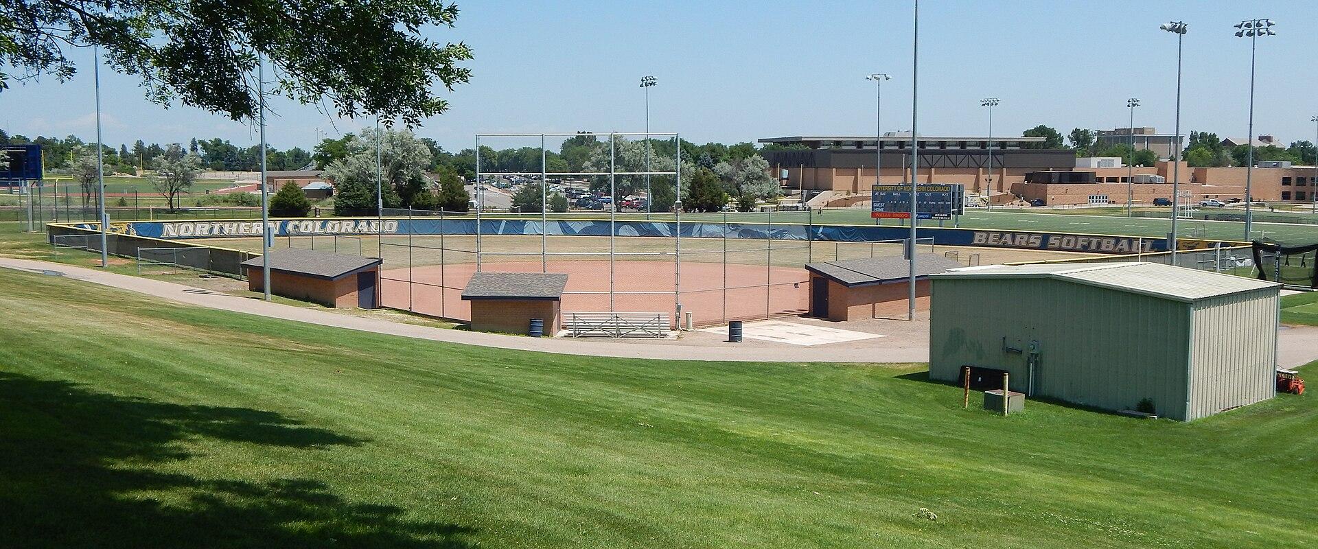 A view of a softball field with a blue outfield fence displaying the text "NORTHERN COLORADO" and "BEARS SOFTBALL", surrounded by green grass and light poles.