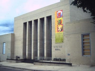 A beige stone building with tall vertical columns and a large vertical banner advertising an Andy Warhol exhibition, with engraved text on the wall reading "COLORADO SPRINGS FINE ARTS CENTER".
