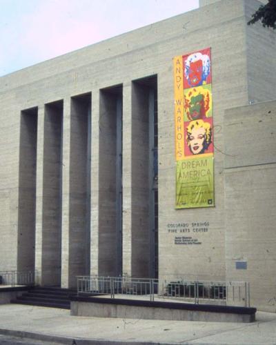 A beige stone building with tall vertical columns and a large vertical banner advertising an Andy Warhol exhibition, with engraved text on the wall reading "COLORADO SPRINGS FINE ARTS CENTER".