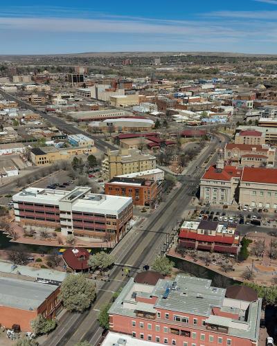 An aerial view of a city center featuring a river with a park and pond, a large red-roofed government building (likely a courthouse), and various brick commercial buildings under a blue sky.